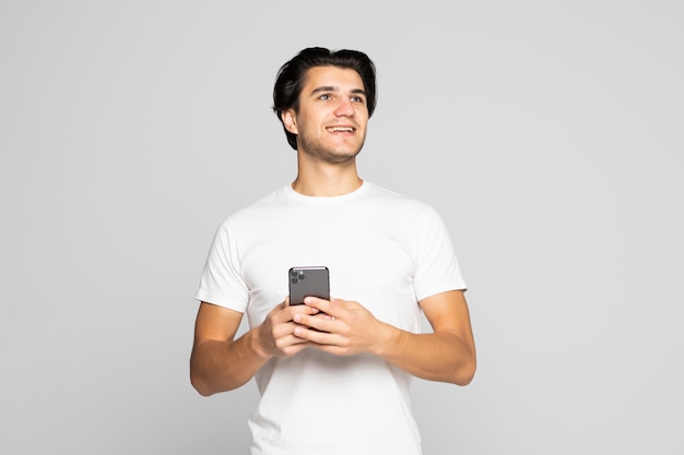 Retrato de luz del día del joven hombre caucásico europeo aislado en gris vestidos de blanco con smartphone