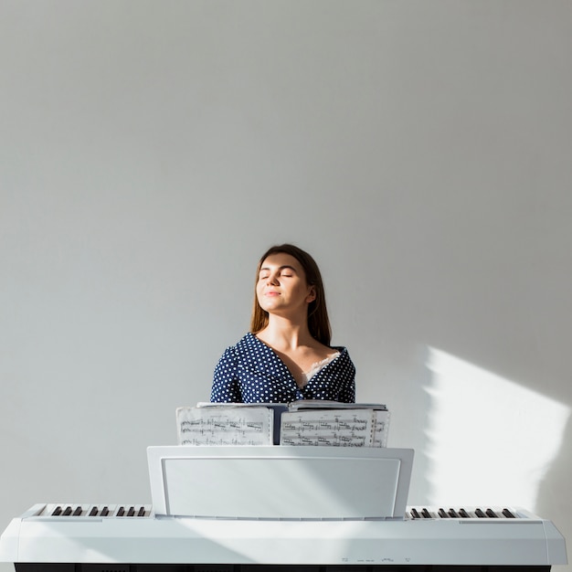 Retrato de una luz del sol de la mujer joven que cierra sus ojos que disfrutan de la luz del sol que se sienta delante de piano