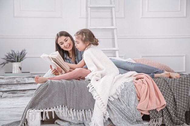 El retrato de una madre y de una hija lindas jovenes sonrientes que leen un libro que miente y se relaja en la cama en un cuarto blanco grande brillante.