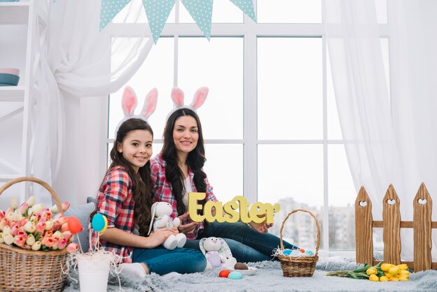 Retrato de la madre y la hija sentada cerca de la ventana con la palabra de Pascua y el conejito en la mano