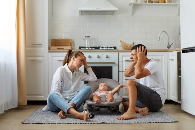 Retrato de madre y padre sentados en el piso de la cocina con el pequeño hijo o hija en una mecedora en el piso de la cocina, los padres cansados y sin dormir manteniendo las manos en la cabeza, necesitan descansar.