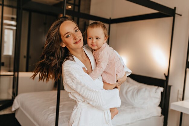 Retrato de madre positiva e hija pequeña en traje de casa posando en el dormitorio. Mujer en bata de baño abrazando al bebé.