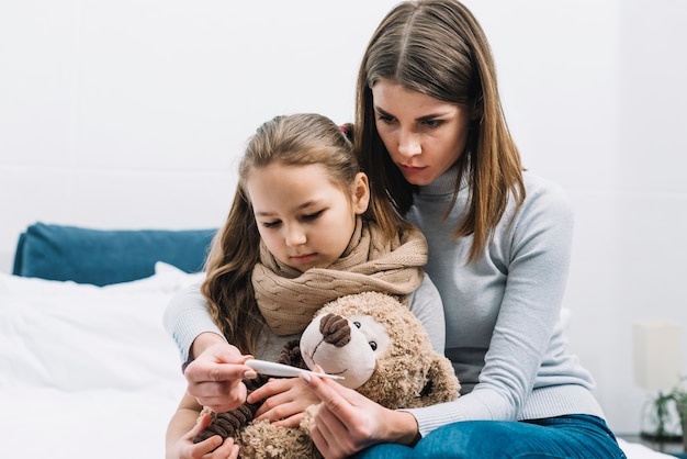 Retrato de la madre sentada con su hija sosteniendo un oso de peluche mirando el termómetro