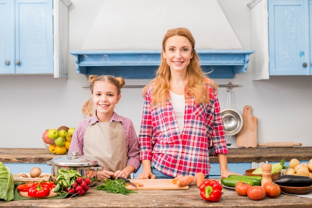 Retrato de una madre sonriente y su hija de pie delante de la mesa con verduras