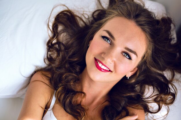 Retrato de mañana de moda tierna de impresionante joven mujer sexy con pecas, pelos esponjosos y maquillaje brillante, acostarse y relajarse en la cama, linda cara sonriente positiva y emociones, colores suaves.