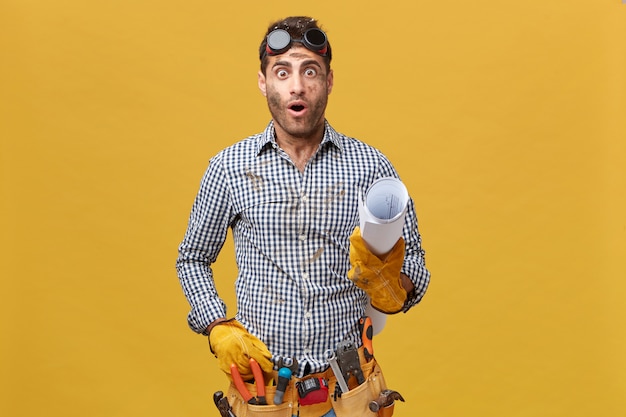 Retrato de manitas sorprendido vistiendo camisa a cuadros, gafas protectoras y guantes, cinturón de herramientas sosteniendo papel enrollado con expresión de sorpresa al darse cuenta de su error. Concepto de personas y trabajo