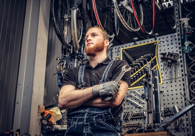 Retrato de un mecánico de bicicletas barbudo con los brazos cruzados sostiene la llave de la taza sobre el fondo del puesto de herramientas en un taller.