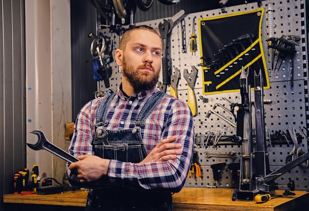 Retrato de un mecánico de bicicletas barbudo con los brazos cruzados sostiene la llave de la taza sobre el fondo del puesto de herramientas en un taller.