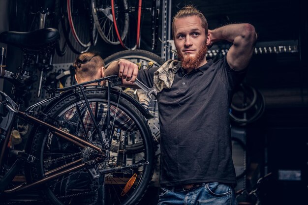 Retrato de un mecánico de bicicletas barbudo de cabeza roja en un taller con piezas de bicicleta y rueda sobre un fondo.