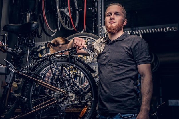 Retrato de un mecánico de bicicletas barbudo de cabeza roja en un taller con piezas de bicicleta y rueda sobre un fondo.