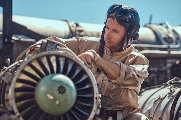 Retrato de un mecánico piloto con uniforme y casco volador, reparando la turbina del avión desmantelada en un museo al aire libre.