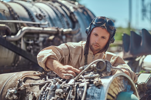 Retrato de un mecánico piloto con uniforme y casco volador, reparando la turbina del avión desmantelada en un museo al aire libre.