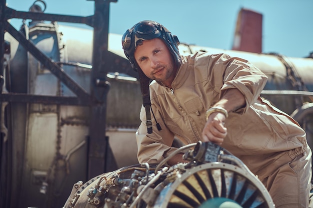 Retrato de un mecánico piloto con uniforme y casco volador, reparando la turbina del avión desmantelada en un museo al aire libre.