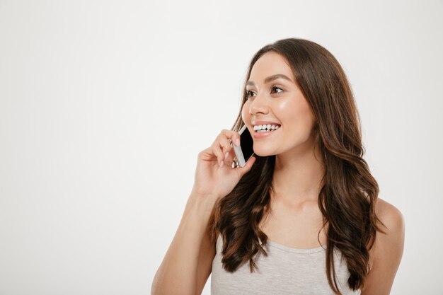Retrato de media vuelta de una mujer caucásica con cabello largo y castaño sonriendo mientras tiene una agradable conversación móvil en su teléfono inteligente, sobre una pared blanca