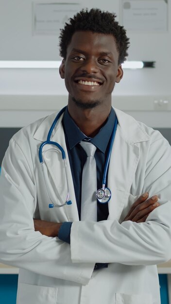 Retrato de un médico afroamericano mirando una cámara con bata blanca y estetoscopio para consultar. Hombre negro sonriendo en el consultorio médico con equipo en el centro de salud