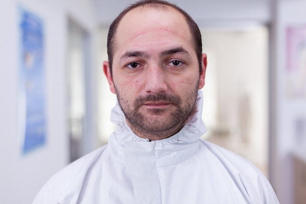 Retrato de médico agotado en la oficina mirando a la cámara con traje de ppe sin protector facial sentado en una silla en la sala de espera