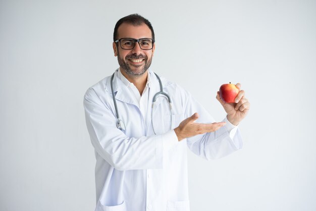 Retrato del médico alegre en los vidrios que ofrecen la manzana madura.