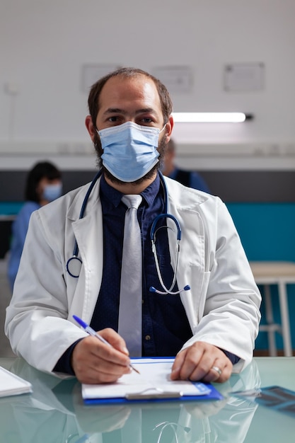 Retrato de un médico con bata blanca y mascarilla en el gabinete, usando papel de portapapeles y bolígrafo para tomar notas en la visita de control. Médico confiado sentado en el escritorio de la oficina durante la pandemia de covid 19.