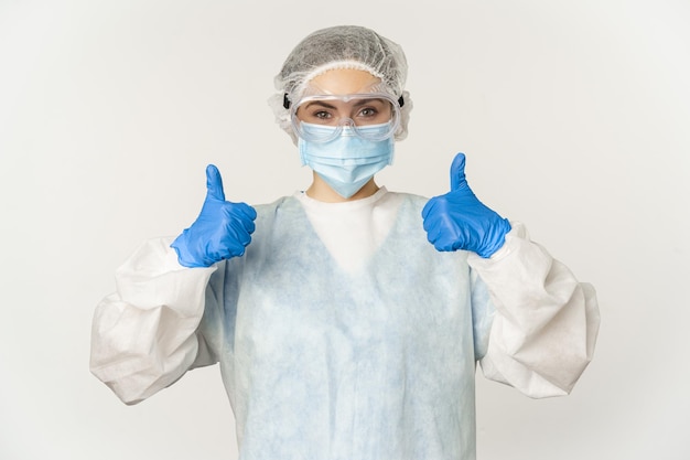 Retrato de médico en equipo de protección personal, mostrando los pulgares hacia arriba, usando máscara facial, concepto de covid-19 y pandemia, fondo blanco.
