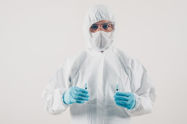 Retrato de un médico en fondo claro con 2 jeringas con medicamentos en guantes médicos y traje protector