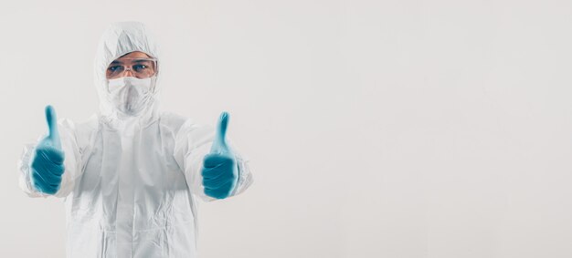 Retrato de un médico en fondo claro de pie y mostrando los pulgares hacia arriba con máscara, guantes médicos y espacio de traje protector para texto