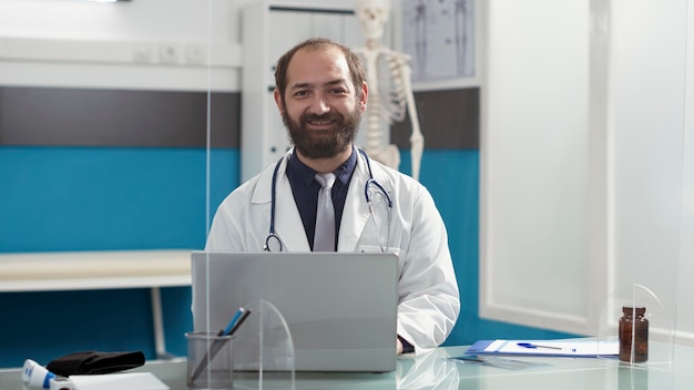 Retrato de un médico general que trabaja en una computadora portátil en el escritorio, usando una computadora para brindar apoyo a los pacientes en la cita de visita de control. Médico masculino con bata blanca y estetoscopio.
