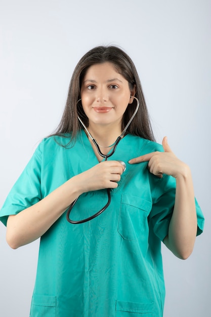 Retrato de un médico joven con estetoscopio en uniforme apuntando a sí misma.