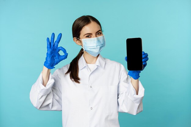 Retrato de un médico con mascarilla médica y guantes que muestra la pantalla del teléfono inteligente de la aplicación de teléfono móvil y está bien...