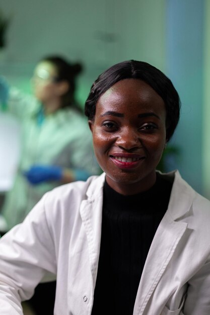Retrato de médico microbiólogo afroamericano mirando a cámara trabajando en el laboratorio del hospital de microbiología. Equipo médico analizando plantas modificadas genéticamente durante un experimento agrícola