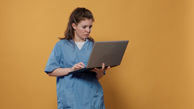 Retrato de un médico ocupado de pie sosteniendo un portátil mirando confundido a la pantalla y escribiendo en el teclado en el estudio. Doctora con uniforme de hospital usando computadora portátil para concepto médico.