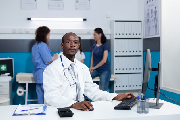 Retrato del médico pediatra afroamericano sentado en la mesa de escritorio en la oficina del hospital