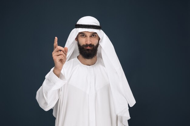 Retrato de medio cuerpo del empresario saudita árabe en la pared azul oscuro. Modelo masculino joven sonriendo y señalando. Concepto de negocio, finanzas, expresión facial, emociones humanas.