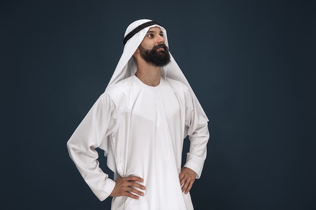 Retrato de medio cuerpo del empresario saudita árabe sobre fondo azul oscuro de estudio. Modelo masculino joven de pie y sonriendo. Concepto de negocio, finanzas, expresión facial, emociones humanas.