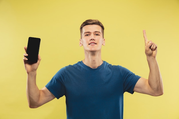 Retrato de medio cuerpo del joven caucásico sobre fondo amarillo de estudio. Hermoso modelo masculino en camisa azul. Concepto de emociones humanas, expresión facial. Mostrando la pantalla del teléfono y apuntando hacia arriba.