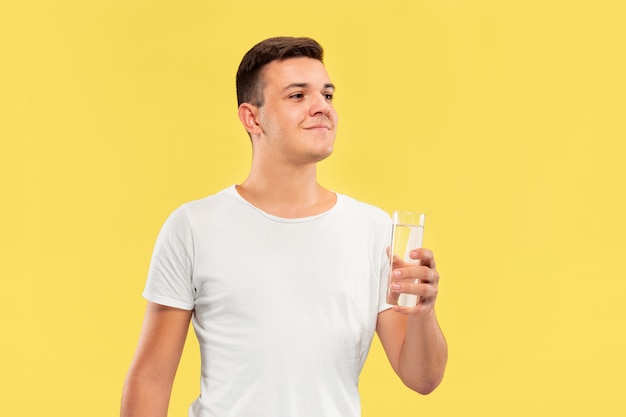 Retrato de medio cuerpo del joven caucásico sobre fondo amarillo de estudio. Hermoso modelo masculino en camisa. Concepto de emociones humanas, expresión facial, ventas, publicidad. Disfrutando bebiendo agua pura.