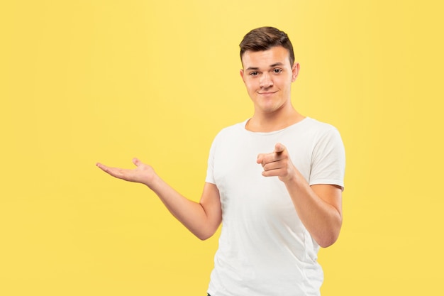 Retrato de medio cuerpo del joven caucásico sobre fondo amarillo de estudio. Hermoso modelo masculino en camisa. Concepto de emociones humanas, expresión facial, ventas, publicidad. Señalar y mostrar algo.