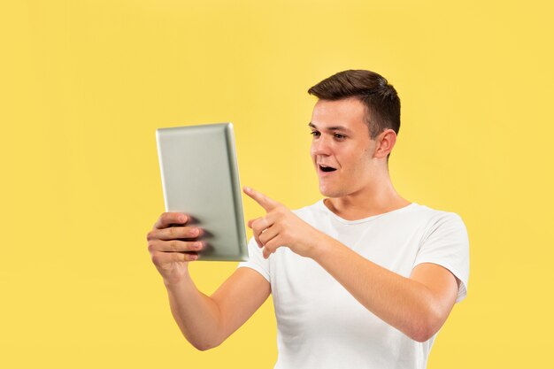 Retrato de medio cuerpo del joven caucásico sobre fondo amarillo de estudio. Hermoso modelo masculino en camisa. Concepto de emociones humanas, expresión facial, ventas, publicidad. Usando tableta y parece feliz.