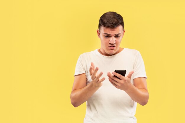 Retrato de medio cuerpo del joven caucásico sobre fondo amarillo de estudio. Hermoso modelo masculino en camisa. Concepto de emociones humanas, expresión facial, ventas, publicidad. Usando el teléfono, se ve sorprendido y molesto.