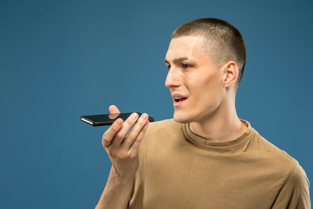 Retrato de medio cuerpo del joven caucásico sobre fondo azul de estudio. Hermoso modelo masculino en camisa. Concepto de emociones humanas, expresión facial, ventas, publicidad. Copyspace. Grabación de mensaje de voz.