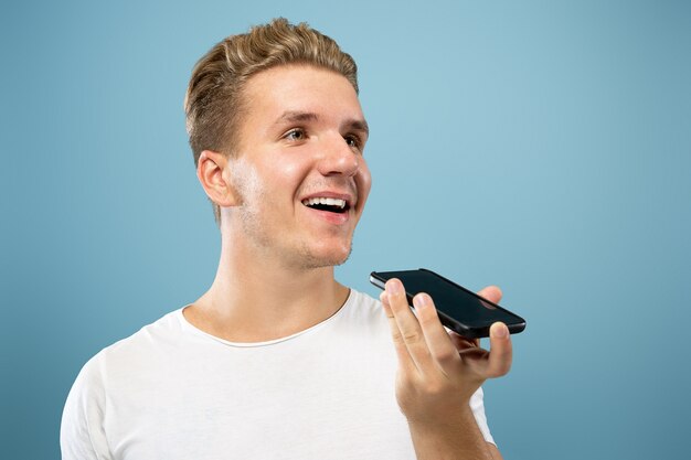 Retrato de medio cuerpo del joven caucásico sobre fondo azul de estudio. Hermoso modelo masculino en camisa. Concepto de emociones humanas, expresión facial, ventas, publicidad. Grabación de mensaje de voz, sonriendo.