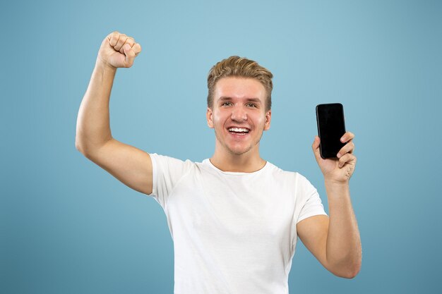 Retrato de medio cuerpo del joven caucásico sobre fondo azul de estudio. Hermoso modelo masculino en camisa. Concepto de emociones humanas, expresión facial, ventas, publicidad. Mostrando la pantalla del teléfono, pago, apuestas.