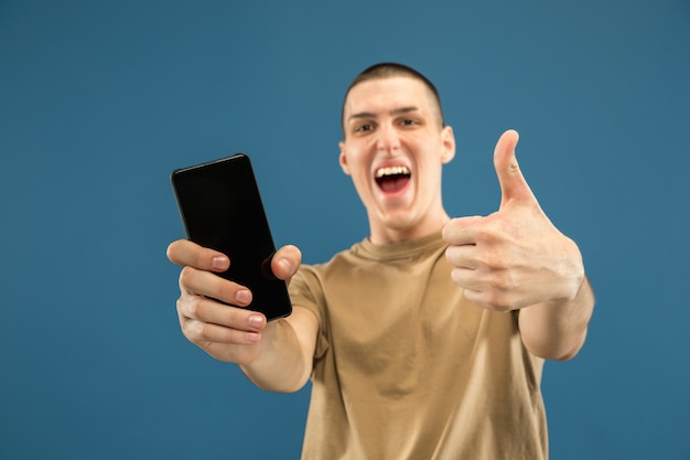 Retrato de medio cuerpo del joven caucásico sobre fondo azul de estudio. Hermoso modelo masculino en camisa. Concepto de emociones humanas, expresión facial, ventas, publicidad. Mostrando la pantalla del teléfono, parece asombrado.