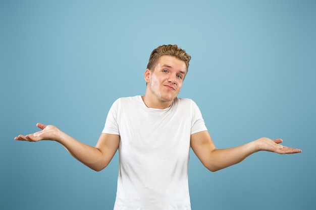 Retrato de medio cuerpo del joven caucásico sobre fondo azul de estudio. Hermoso modelo masculino en camisa. Concepto de emociones humanas, expresión facial, ventas, publicidad. Mostrando, parece incierto, copyspace.