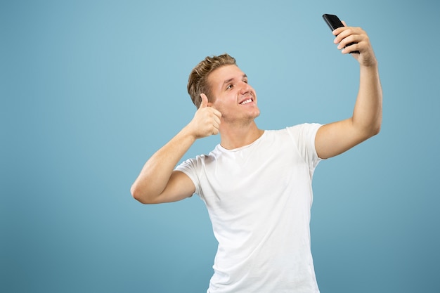 Retrato de medio cuerpo del joven caucásico sobre fondo azul de estudio. Hermoso modelo masculino en camisa. Concepto de emociones humanas, expresión facial, ventas, publicidad. Realización de selfies o contenido para vlog.