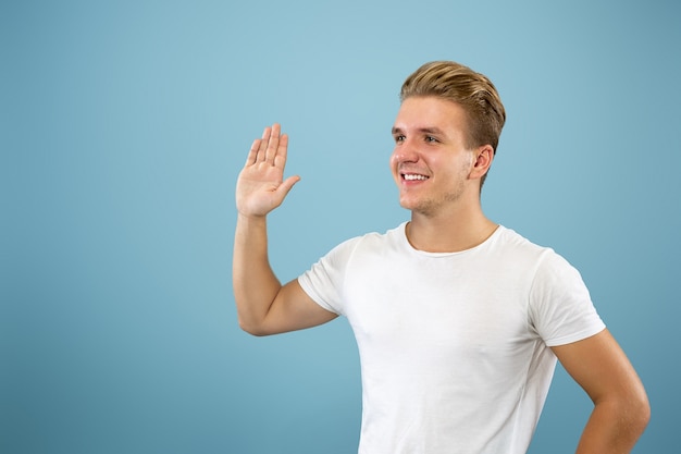 Retrato de medio cuerpo del joven caucásico sobre fondo azul de estudio. Hermoso modelo masculino en camisa. Concepto de emociones humanas, expresión facial, ventas, publicidad. Saludar, invitar a alguien.