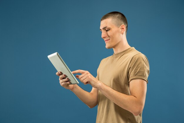 Retrato de medio cuerpo del joven caucásico sobre fondo azul de estudio. Hermoso modelo masculino en camisa. Concepto de emociones humanas, expresión facial, ventas, publicidad. Usando tableta para selfie, vlog o estudio.