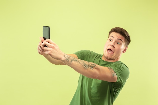 Retrato de medio cuerpo del joven caucásico sobre fondo verde de estudio. Hermoso modelo masculino en camisa. Concepto de emociones humanas, expresión facial, ventas, publicidad. Haciendo selfie o contenido para su vlog.