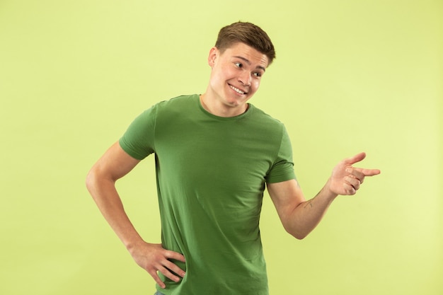 Retrato de medio cuerpo del joven caucásico sobre fondo verde de estudio. Hermoso modelo masculino en camisa. Concepto de emociones humanas, expresión facial, ventas, publicidad. Señalando, parece confiado.