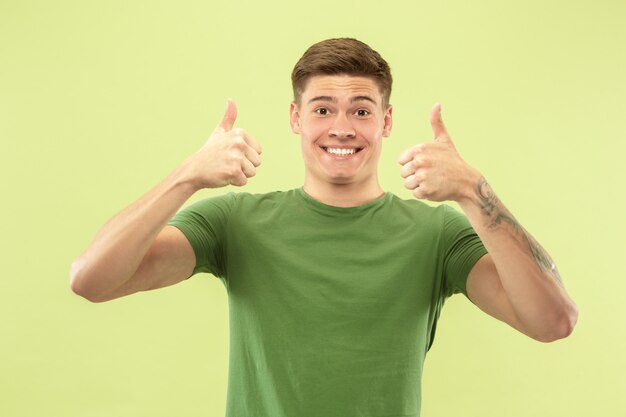 Retrato de medio cuerpo del joven caucásico sobre fondo verde de estudio. Hermoso modelo masculino en camisa. Concepto de emociones humanas, expresión facial, ventas, publicidad. Sonriendo, mostrando los pulgares hacia arriba.