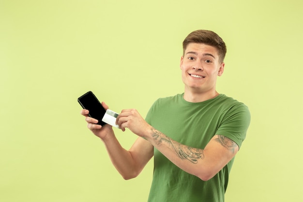 Retrato de medio cuerpo del joven caucásico sobre fondo verde de estudio. Hermoso modelo masculino en camisa. Concepto de emociones humanas, expresión facial, ventas, publicidad. Sosteniendo teléfono y tarjeta, pagos en línea.